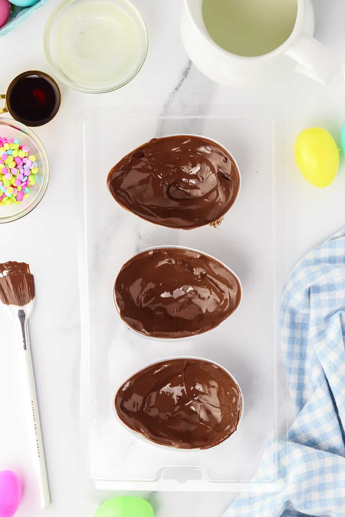 Three chocolate Easter egg halves in a mold, surrounded by ingredients and tools for making homemade Copycat Creme Egg candy.
