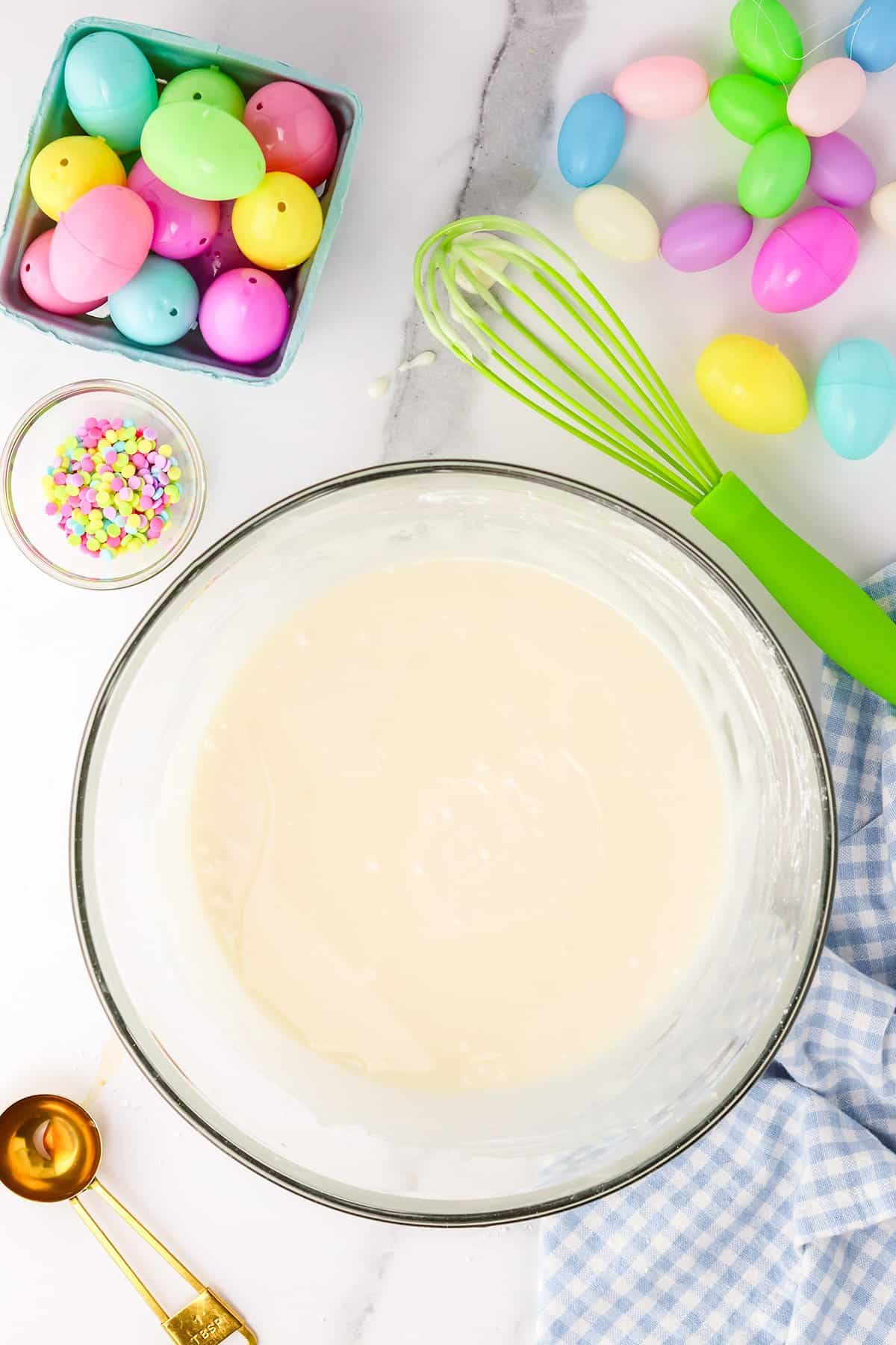 Ingredients and utensils for Easter baking, featuring colorful plastic eggs, Creme Egg Candy, and cake batter on a kitchen countertop.