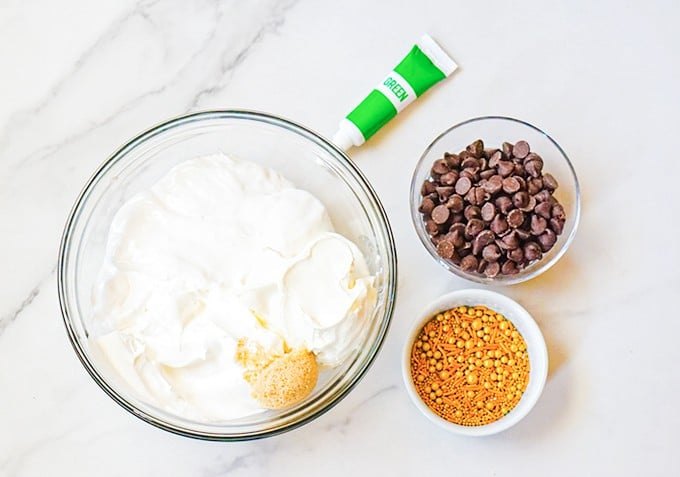 Ingredients for a St. Patrick's Day dessert dip on a marble countertop: a bowl of whipped cream with chocolate chips and sprinkles