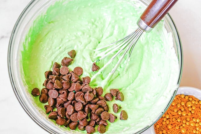 A bowl of green dip being mixed with a whisk for St. Patrick's Day, with chocolate chips added on top.