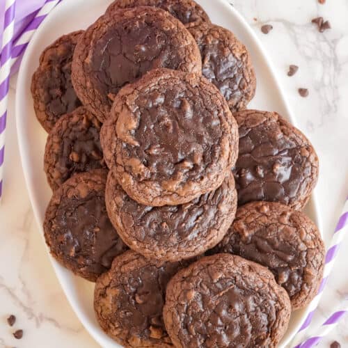 A stack of brownie mix cookies on a white marble countertop, accompanied by purple striped straws and scattered chocolate chips.