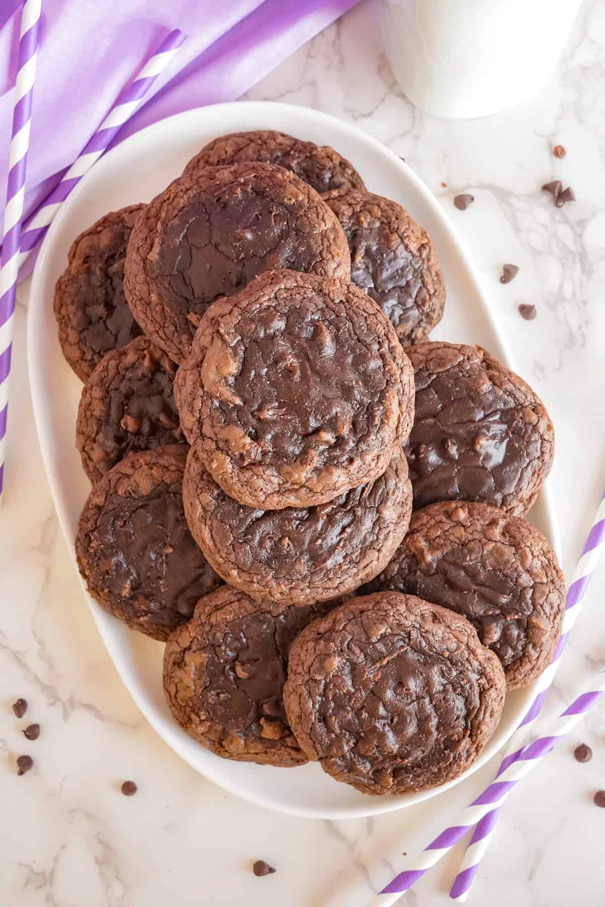 A stack of brownie mix cookies on a white marble countertop, accompanied by purple striped straws and scattered chocolate chips.