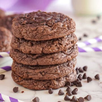 A stack of four brownie mix cookies with chocolate chips scattered around, on a marble surface with a striped purple and white background.