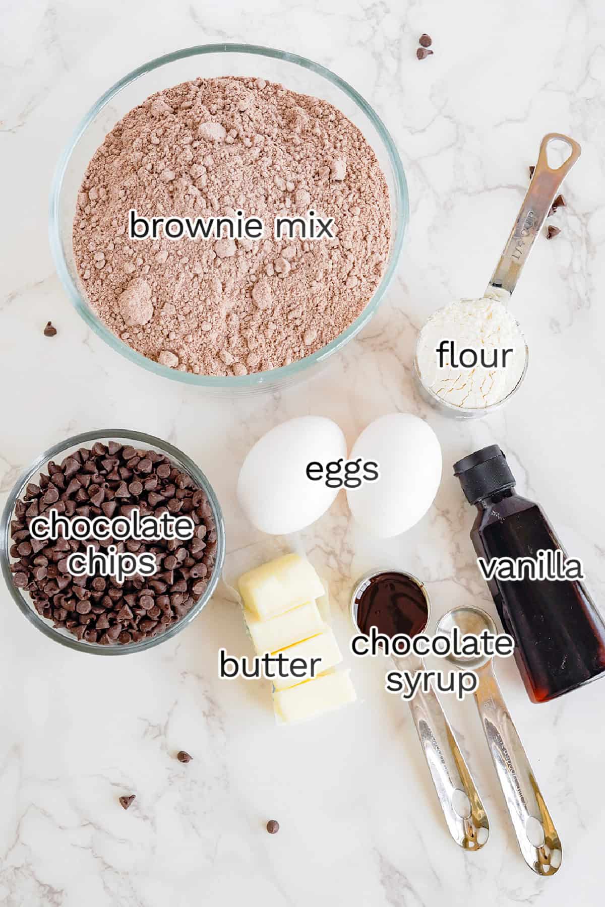 Baking ingredients on a marble countertop including brownie mix cookies, flour, chocolate chips, eggs, butter, and chocolate syrup labeled for clarity.