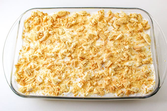 A glass dish filled with a lemon delight dessert, topped with crushed golden cookies, sits on a white background.