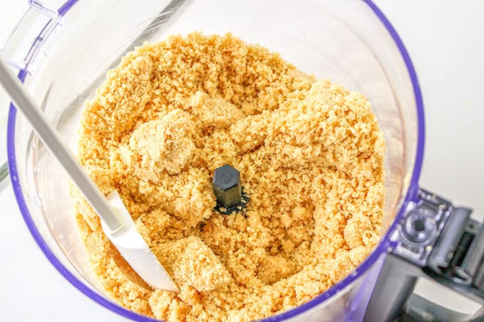 Crushed graham crackers in a food processor with a spatula, viewed from above, creating the perfect base for a refreshing lemon delight.