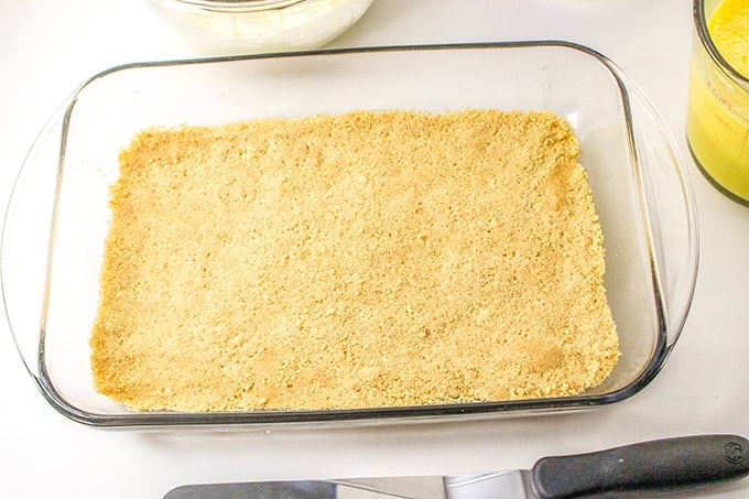 A glass baking dish is filled with a layer of finely ground yellow crumbs, likely from graham crackers or a similar ingredient, hinting at the start of a lemon delight. A knife and a glass of liquid are placed nearby.