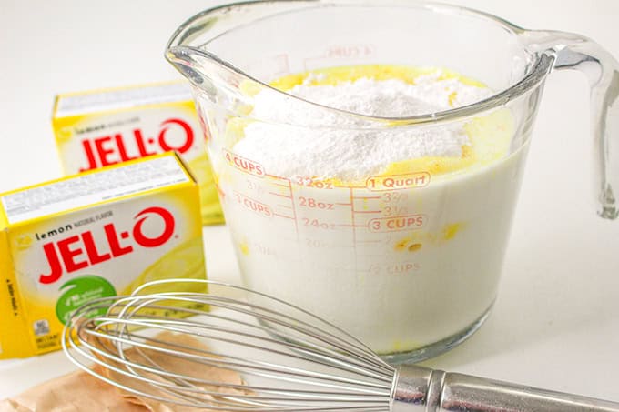 A glass measuring cup filled with milk, butter, and powder mixture sits beside two boxes of lemon Jell-O and a metal whisk, ready to create a lemon delight.