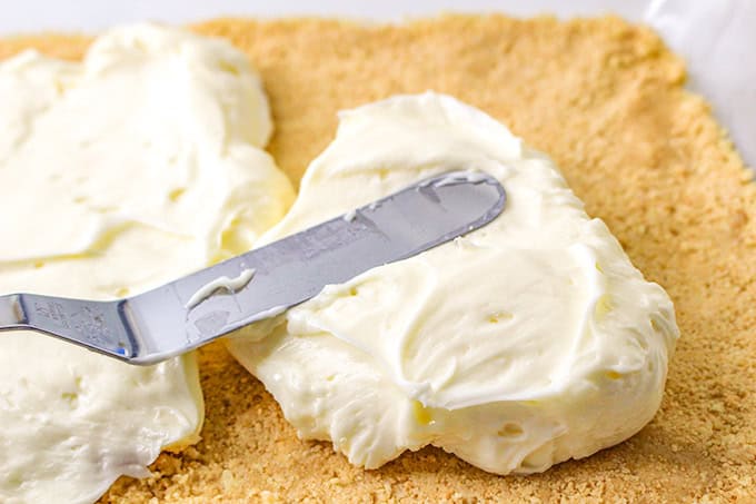 A metal spatula spreads lemon delight cream cheese filling over a graham cracker crust.