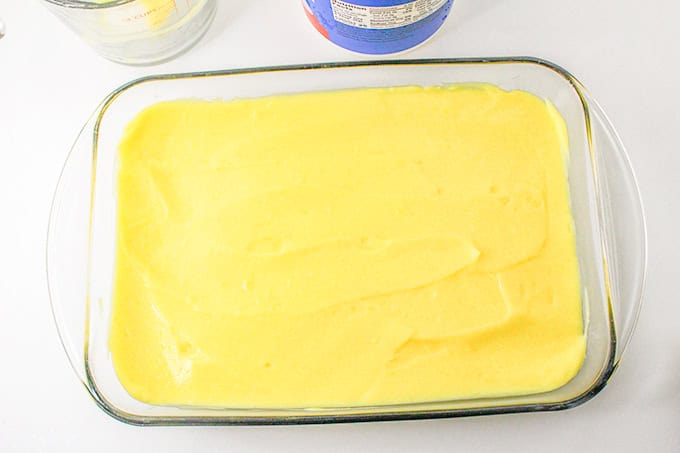 A rectangular glass baking dish filled with a smooth layer of yellow lemon delight batter rests on a white surface, accompanied by partially visible containers.