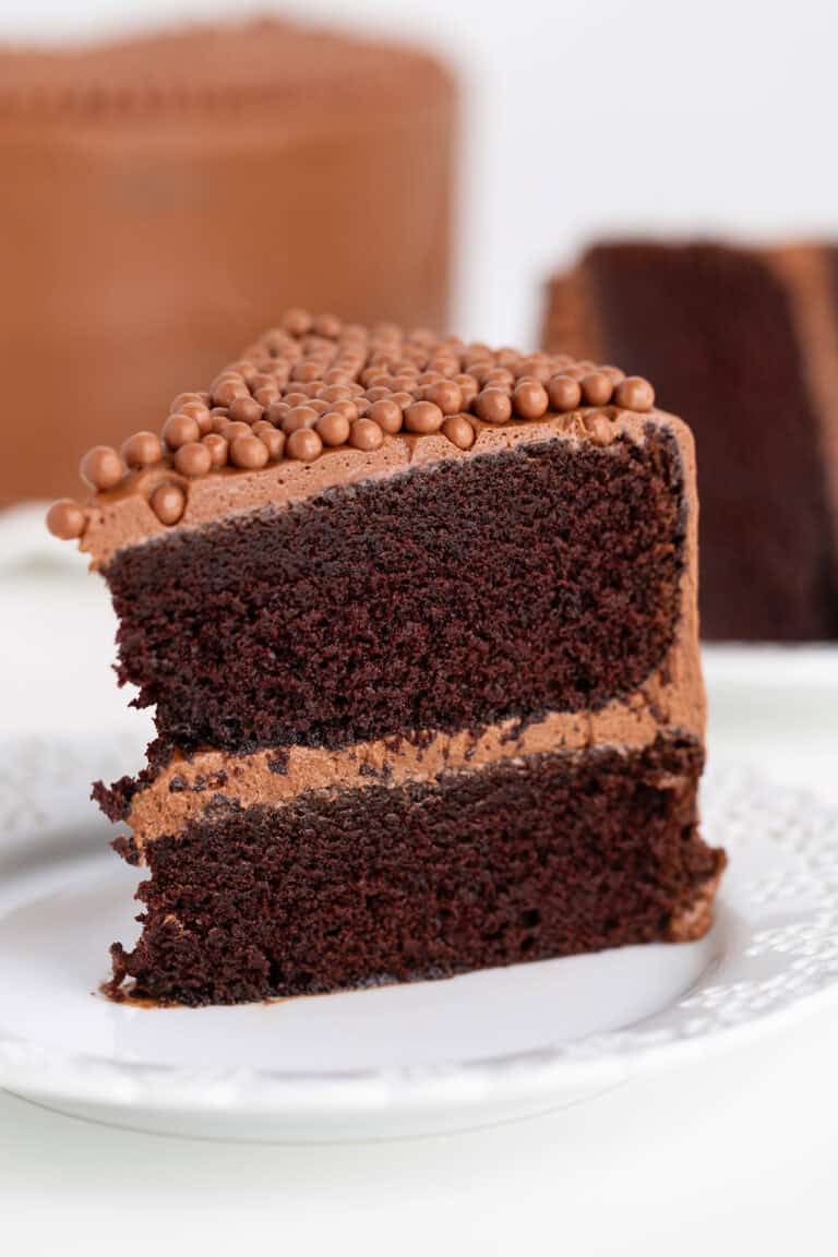 A slice of Ina Garten's Chocolate Cake with two layers of frosting and chocolate pearls on top, served on a white plate with more cake in the background.