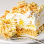 Close-up of a plate with a slice of layered lemon delight dessert, including crumbled topping, whipped cream, a zesty yellow filling, and a crumbly base. A fork rests on the plate beside the dessert.