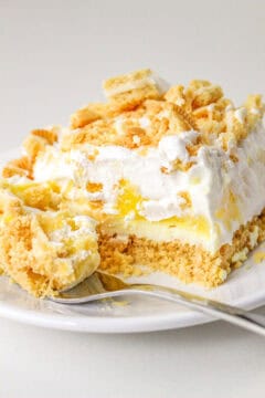 Close-up of a plate with a slice of layered lemon delight dessert, including crumbled topping, whipped cream, a zesty yellow filling, and a crumbly base. A fork rests on the plate beside the dessert.