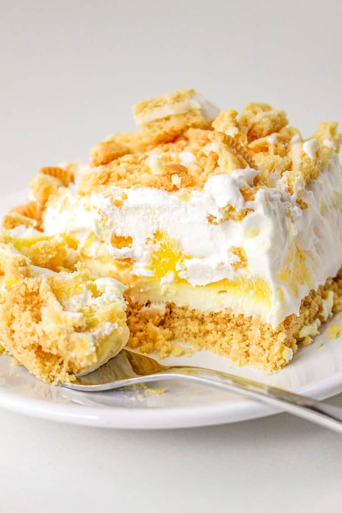 Close-up of a plate with a slice of layered lemon delight dessert, including crumbled topping, whipped cream, a zesty yellow filling, and a crumbly base. A fork rests on the plate beside the dessert.