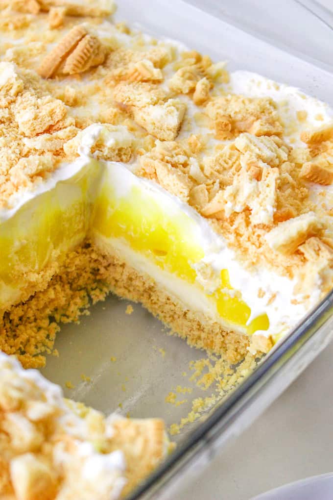 A dessert in a glass dish with a cut section revealing layers of crushed cookies, creamy filling, and a bright yellow layer of lemon delight, topped with crumbly cookie pieces.