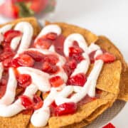 A plate of nachos topped with white sauce and a strawberry mixture, resembling a delightful cheesecake, with a bowl of fresh strawberries in the background on a white surface with a red polka dot cloth underneath.