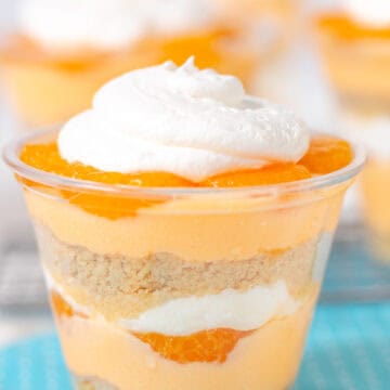 A layered dessert cup reminiscent of a creamsicle, with whipped cream on top, featuring layers of orange segments, cream, and cookie crumbs, all placed on a blue polka-dotted napkin.