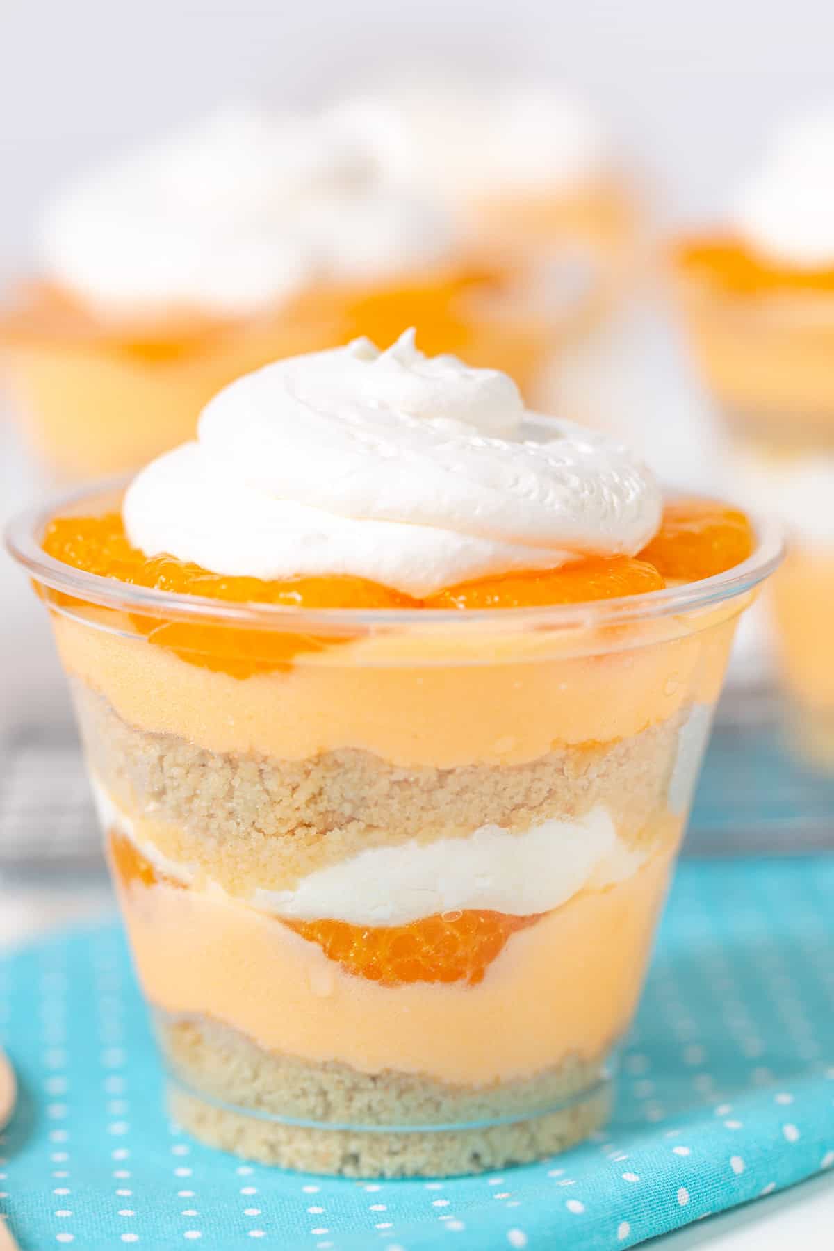 A layered dessert cup reminiscent of a creamsicle, with whipped cream on top, featuring layers of orange segments, cream, and cookie crumbs, all placed on a blue polka-dotted napkin.
