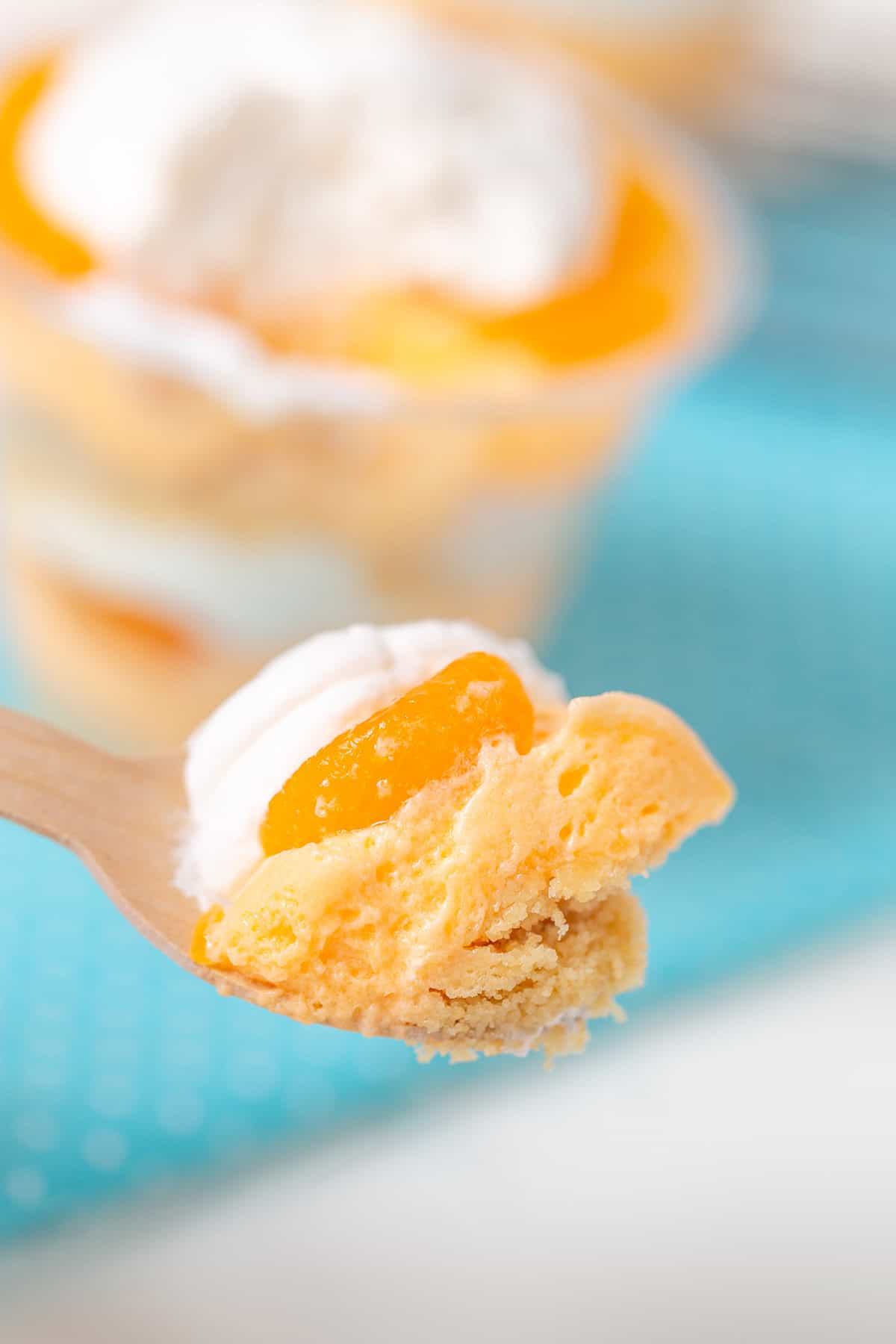 A close-up of a spoon holding a bite of creamsicle dessert with a piece of orange fruit and whipped cream. The background is blurred, featuring multiple cups filled with desserts on a blue surface.