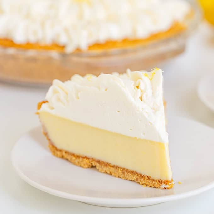 A slice of lemon pie with a graham cracker crust, creamy lemon filling, and a whipped topping sits invitingly on a white plate.