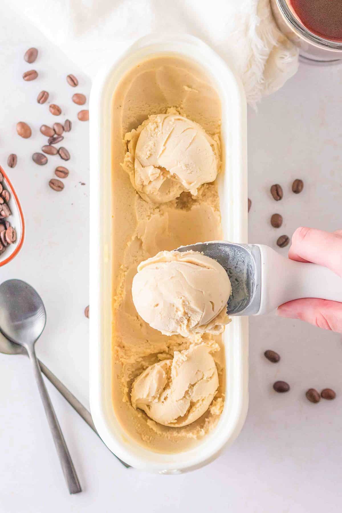 Hand scooping coffee ice cream from a rectangular container with coffee beans scattered around and a spoon placed beside it, this delightful indulgence belongs to the Ice Cream category.