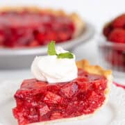 A slice of strawberry pie topped with whipped cream and a mint leaf sits on a white plate, with the remaining strawberry pie and a bowl of strawberries in the background.