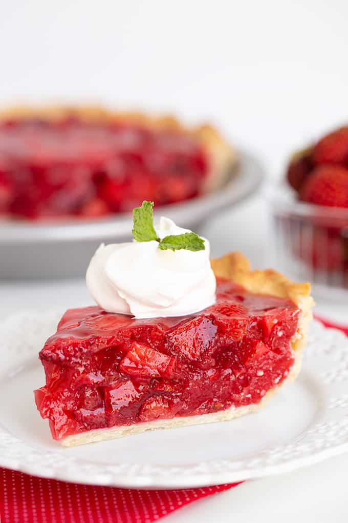 A slice of strawberry pie topped with whipped cream and a mint leaf sits on a white plate, with the remaining strawberry pie and a bowl of strawberries in the background.