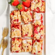 A rectangular white plate holds strawberry rhubarb bars drizzled with glaze. Two whole strawberries are placed at the top. Two gold forks and two empty plates are beside the dish.