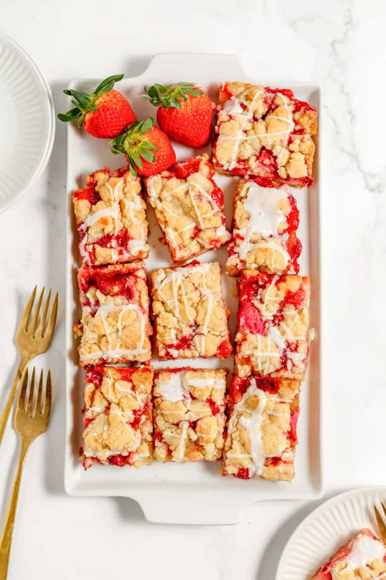 A rectangular white plate holds strawberry rhubarb bars drizzled with glaze. Two whole strawberries are placed at the top. Two gold forks and two empty plates are beside the dish.
