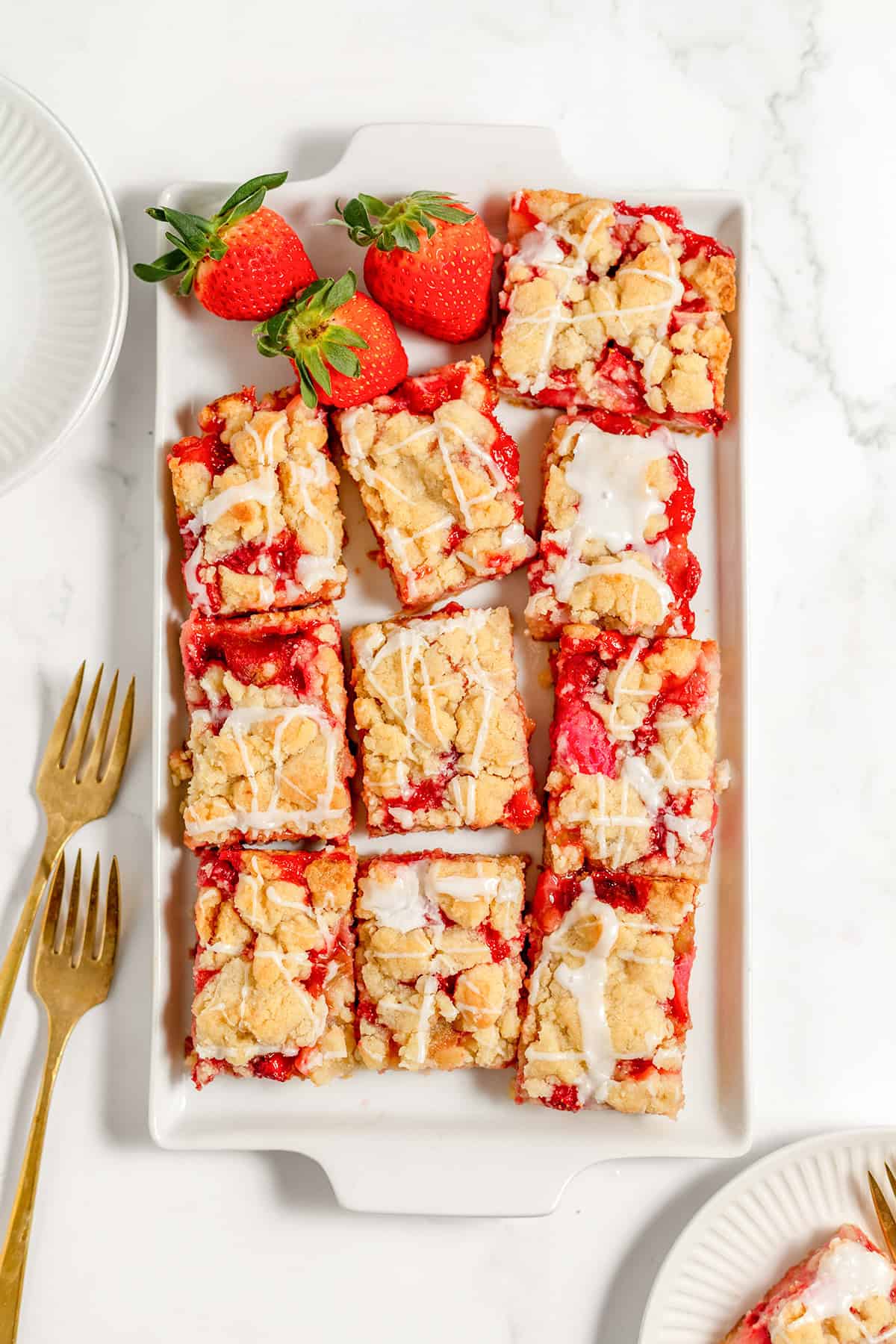 A rectangular white plate holds strawberry rhubarb bars drizzled with glaze. Two whole strawberries are placed at the top. Two gold forks and two empty plates are beside the dish.