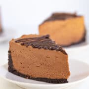 Slice of no-bake chocolate cheesecake with a chocolate drizzle topping on a white plate, against a plain white background.