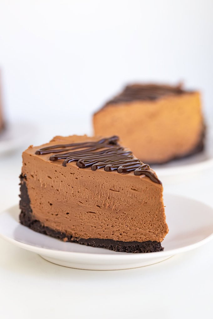 Slice of no-bake chocolate cheesecake with a chocolate drizzle topping on a white plate, against a plain white background.
