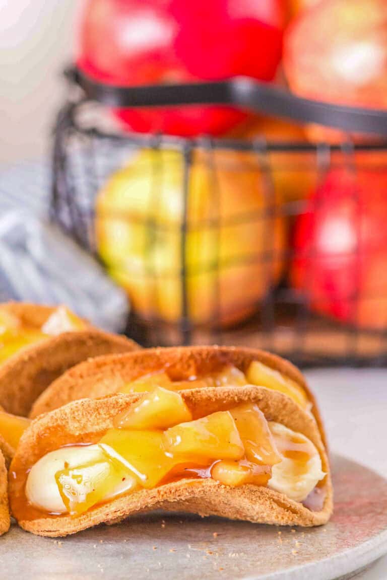 Close-up of Apple Cheesecake Tacos filled with caramelized fruit and cream, placed on a gray plate. A wire basket containing apples and a wrapped item is in the blurred background.