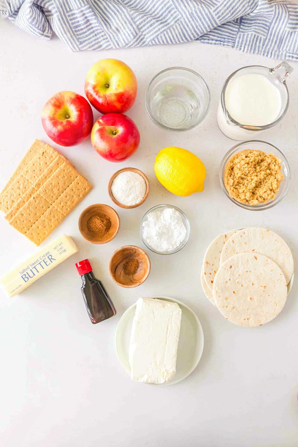 Ingredients on a countertop including apples, graham crackers, lemon, butter, brown sugar, flour, tortillas for delicious Apple Cheesecake Tacos, vanilla extract, cinnamon, baking powder, salt, water, and milk.