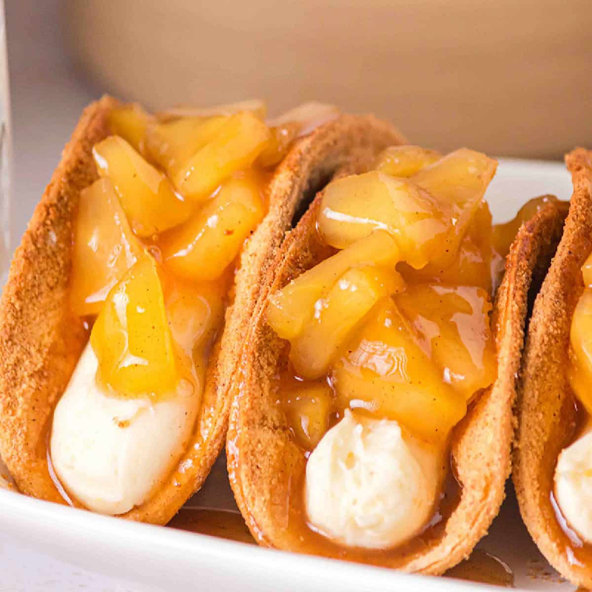 Three dessert tacos filled with whipped cream and topped with a caramelized apple and cinnamon mixture on a white plate, reminiscent of a deconstructed cheesecake.