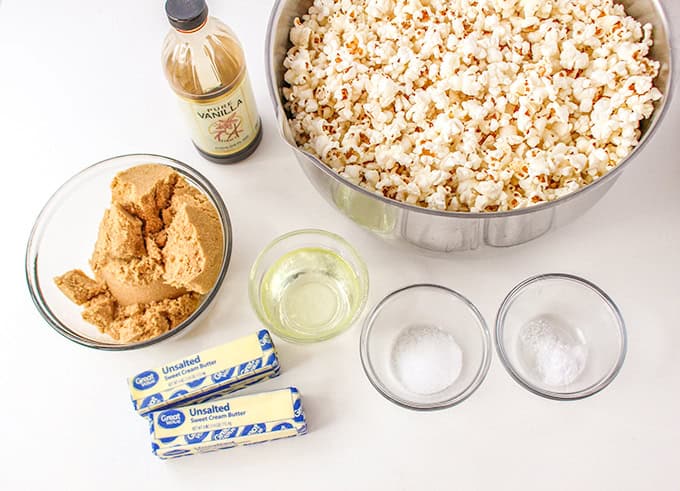 A large bowl of old-fashioned caramel corn sits next to ingredients on the countertop, including brown sugar, vanilla extract, unsalted butter, vegetable oil, salt, and baking soda in separate bowls.
