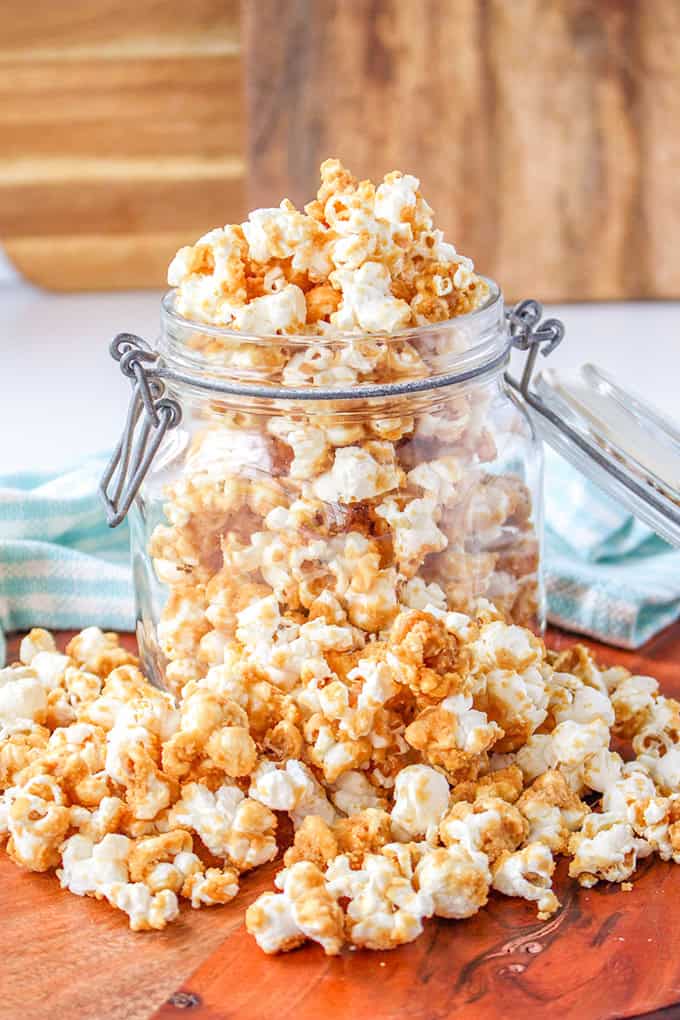 An old-fashioned glass jar filled with caramel corn is open, with some popcorn spilling out onto a wooden surface. A striped cloth is visible in the background.