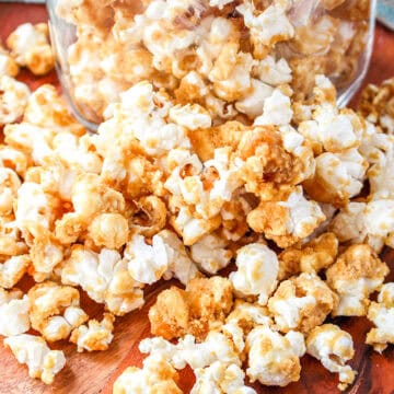 An old-fashioned glass jar filled with caramel corn sits on a wooden surface, with some popcorn charmingly spilled out around it.