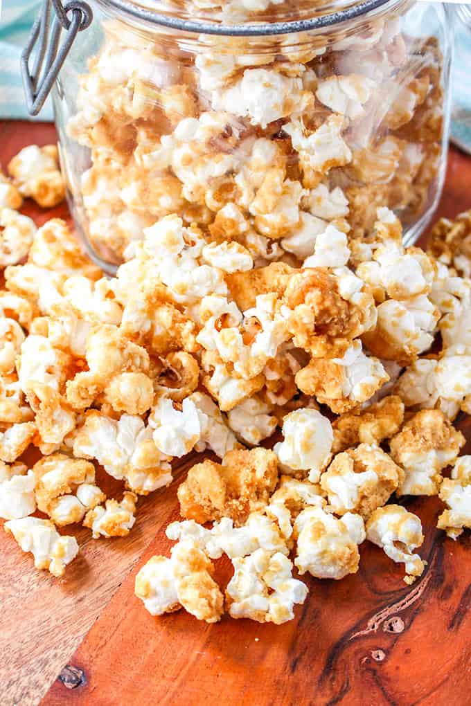 An old-fashioned glass jar filled with caramel corn sits on a wooden surface, with some popcorn charmingly spilled out around it.