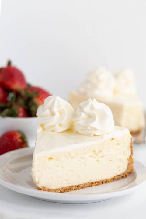 A slice of homemade cheesecake topped with two dollops of whipped cream sits on a plate, while a bowl of fresh strawberries is blurred in the background.