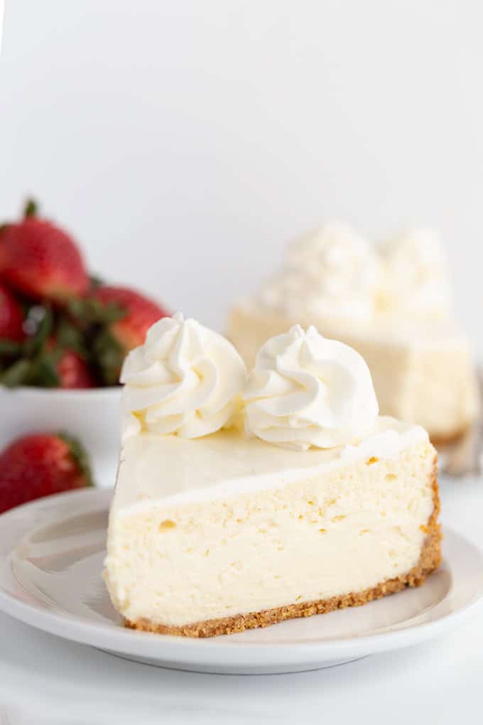 A slice of homemade cheesecake topped with two dollops of whipped cream sits on a plate, while a bowl of fresh strawberries is blurred in the background.