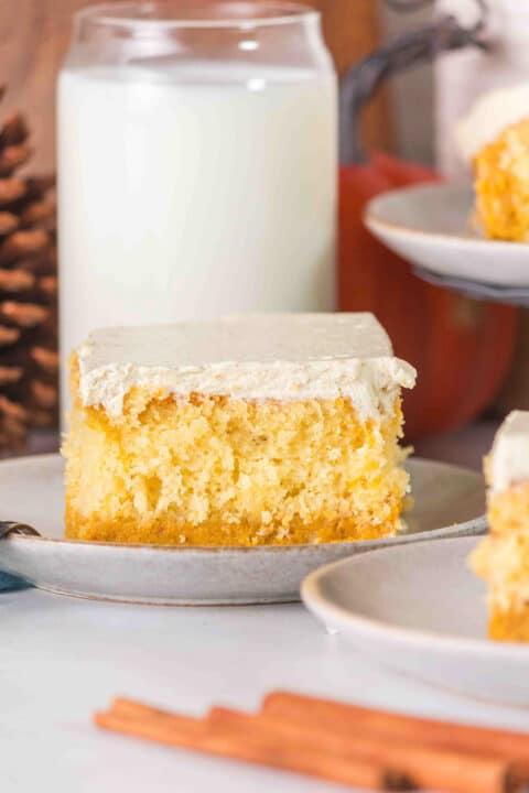 A slice of pumpkin magic cake with white frosting sits on a plate, paired with a glass of milk and another plate of cake in the background.