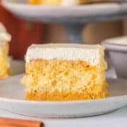 A slice of pumpkin magic cake with a thick layer of white frosting is served on a white plate, with cinnamon sticks nearby.