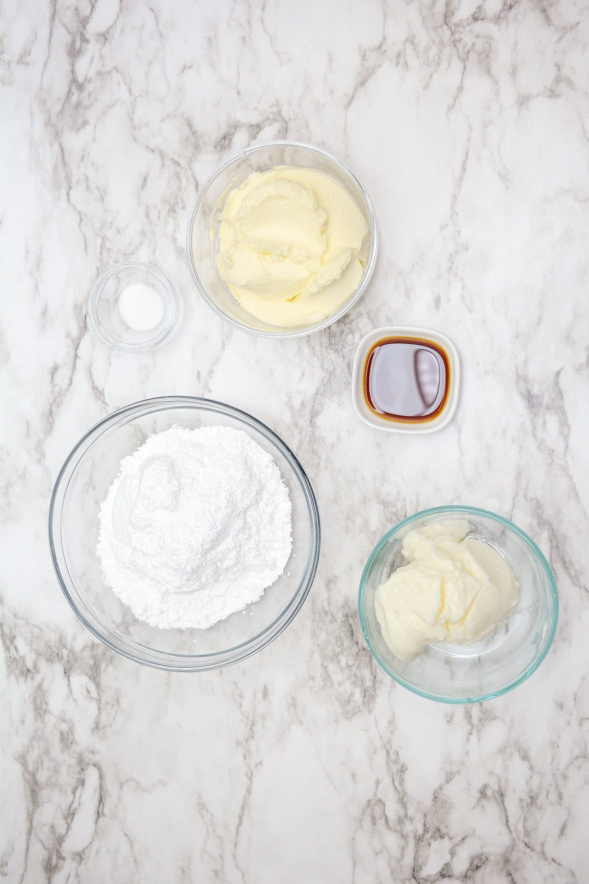 Ingredients for frosting on a marble surface: a bowl of butter, a bowl of powdered sugar, and a bowl of shortening await their transformation into the perfect topping for your pumpkin roll. Add in a small bowl of vanilla extract and a pinch of salt to complete this cake-worthy creation.