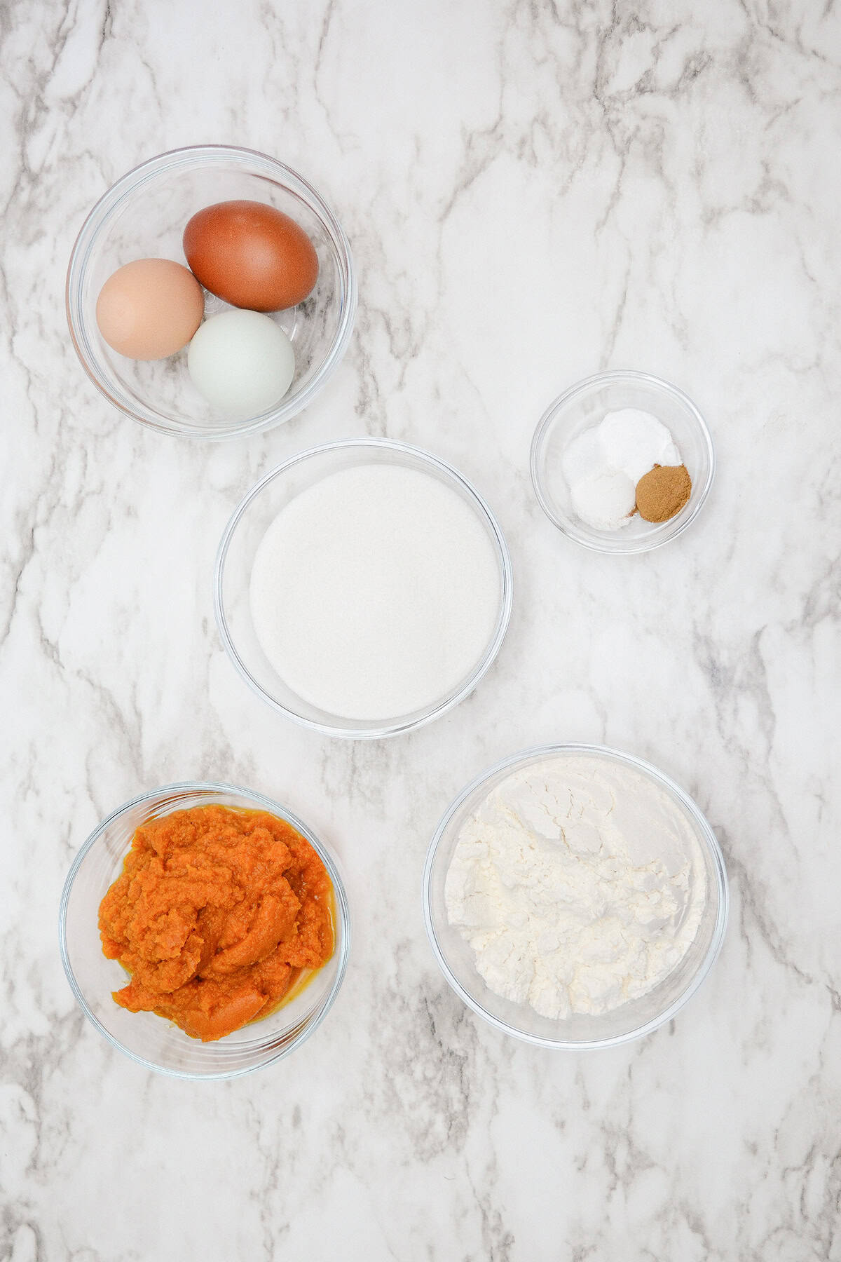 Baking ingredients on a marble countertop: two eggs, sugar, flour, and pumpkin puree sit ready. Nearby, small bowls of salt and cinnamon add a touch of spice to the pumpkin cake waiting to roll into life.