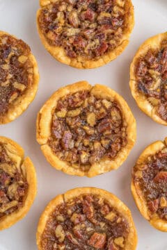 A plate of six mini pecan pies arranged in a circular pattern, featuring a flaky crust and nutty filling.
