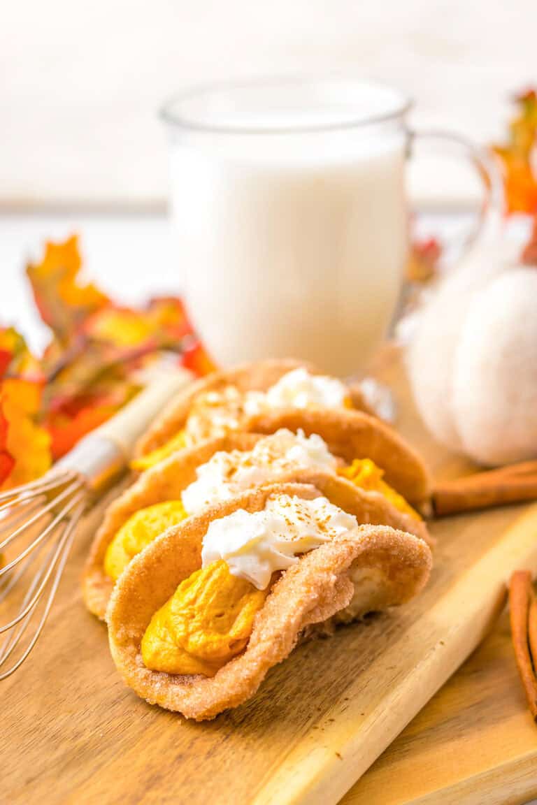 Three pumpkin pie-inspired dessert tacos filled with creamy pumpkin and topped with fluffy whipped cream rest on a wooden board. A glass of milk sits in the background, surrounded by autumn leaves and a whisk.