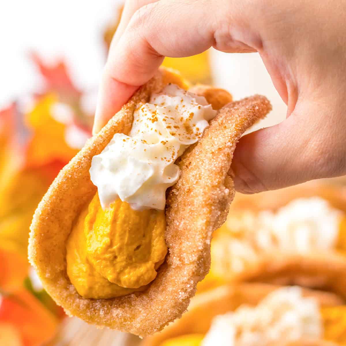 A hand holds a dessert taco reminiscent of pumpkin pie, filled with silky pumpkin mousse, topped with whipped cream and a dusting of cinnamon, all set against a backdrop of colorful autumn leaves.