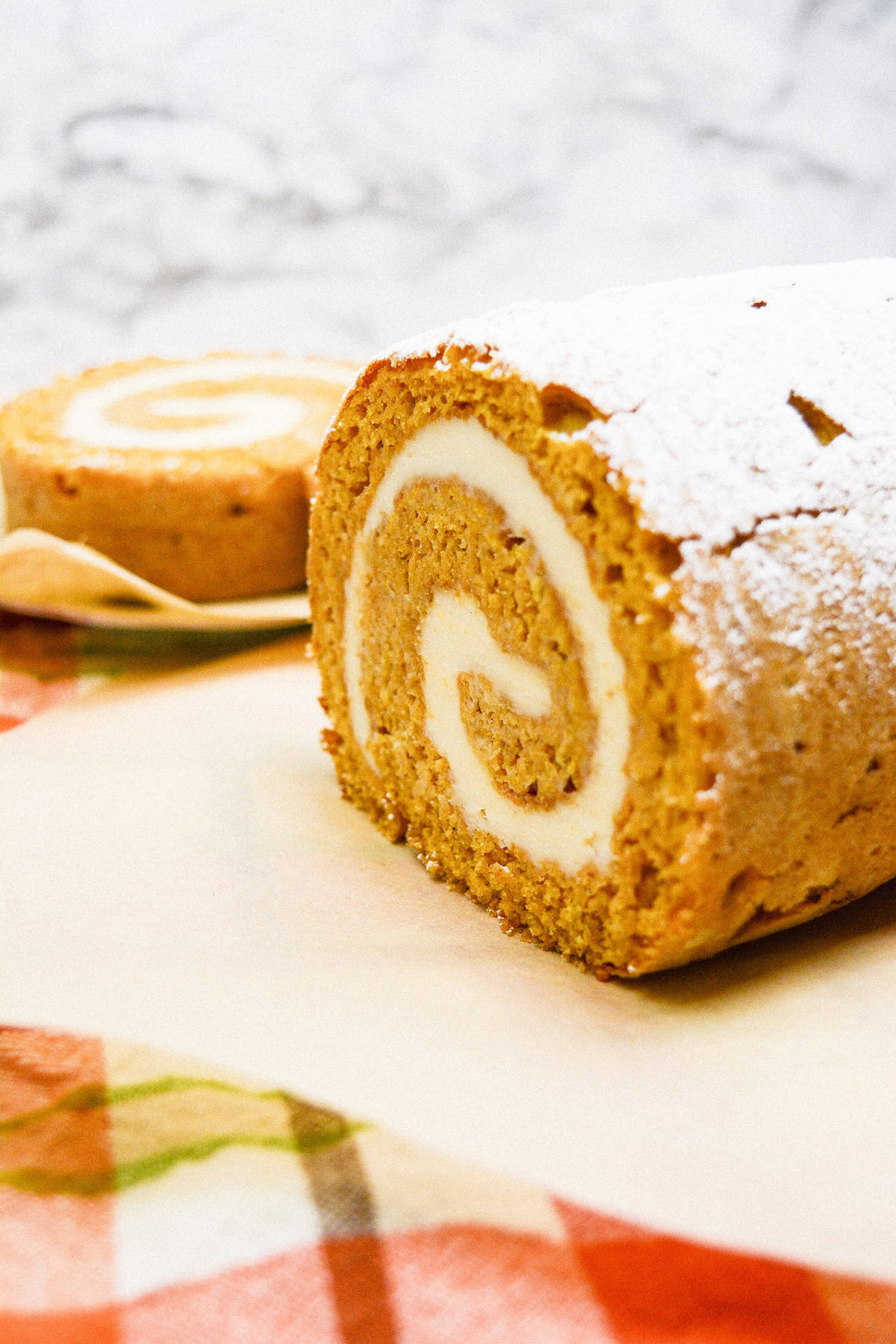 A delicious pumpkin roll cake with cream cheese filling, dusted with powdered sugar, graces a table adorned with a checkered cloth. In the background, a tempting slice of this pumpkin roll awaits on a plate.