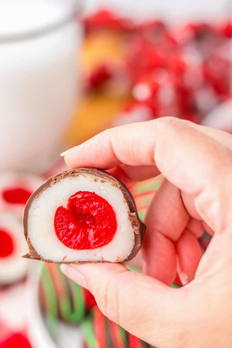 A hand holds a chocolate-covered cherry cordial, revealing a glossy white filling with a vibrant red center.